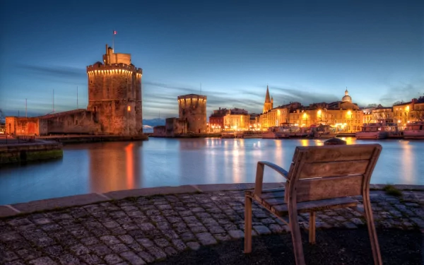 HD desktop wallpaper showcasing a man-made town at dusk with historic towers, waterfront buildings, and a wooden bench in the foreground.