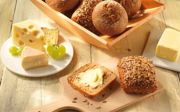 HD desktop wallpaper showing an assortment of bread rolls, slices of cheese, butter, and grapes arranged on a wooden table.