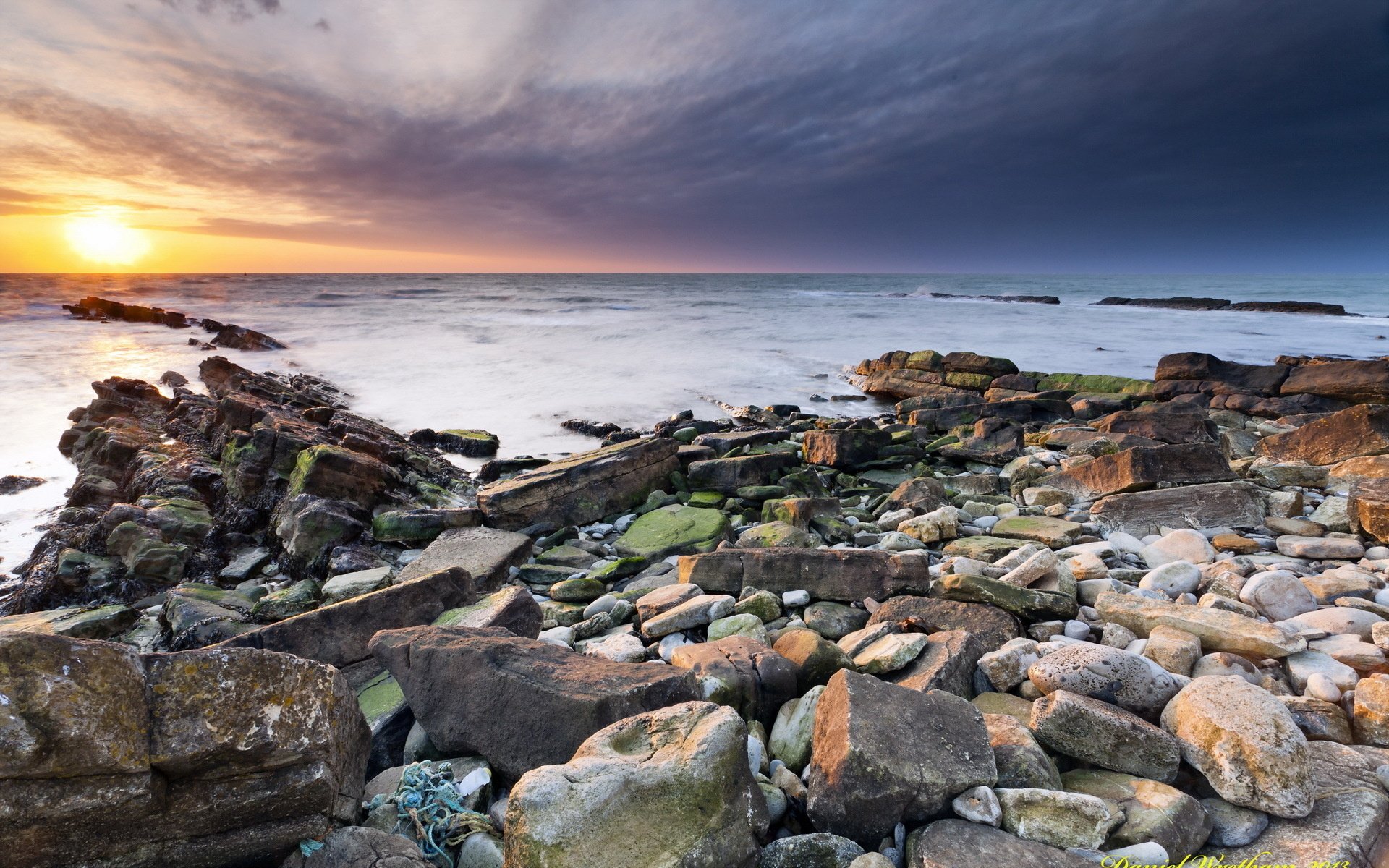 HD desktop wallpaper showcasing a rocky shoreline at sunset with waves gently washing over the stones under a vibrant sky.