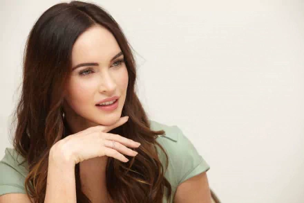 Megan Fox, featured in this 4K Ultra HD desktop wallpaper, showcases a thoughtful expression with her hand resting on her chin, framed by her flowing hair.