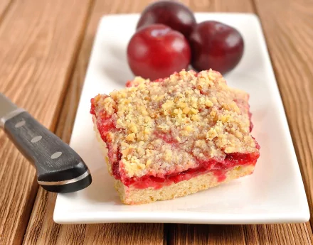 HD desktop wallpaper showing a piece of crumb-topped fruit bar with red filling, served on a white plate alongside two plums and a knife, highlighting food and baking.