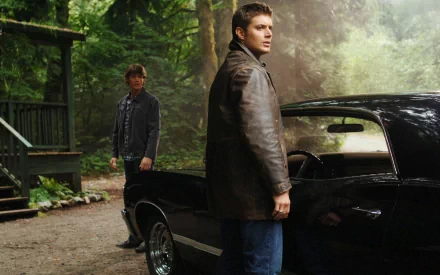 Two main characters from the TV show Supernatural stand in a forested area, one in a leather jacket, next to a classic black car, capturing a dramatic moment in HD wallpaper format.