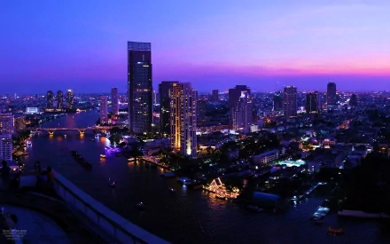 HD desktop wallpaper showcasing the vibrant Bangkok skyline at twilight with illuminated skyscrapers and the winding river below.