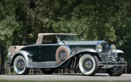 A 1929 Duesenberg Model J convertible parked on a driveway with lush greenery in the background, showcasing its classic design and luxurious details.