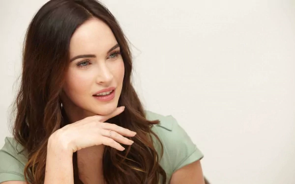 Megan Fox, featured in this 4K Ultra HD desktop wallpaper, showcases a thoughtful expression with her hand resting on her chin, framed by her flowing hair.