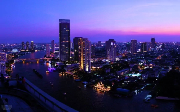 HD desktop wallpaper showcasing the vibrant Bangkok skyline at twilight with illuminated skyscrapers and the winding river below.