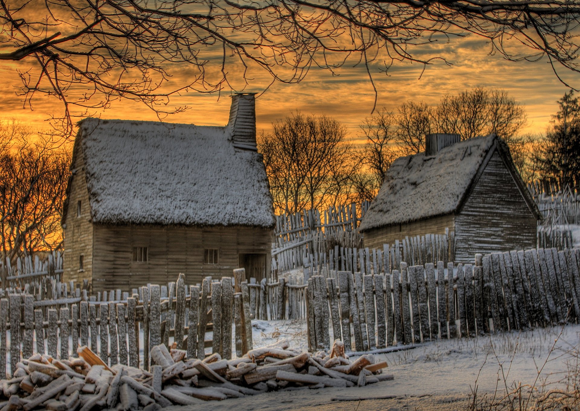 HD PC desktop wallpaper featuring a man-made rustic cabin with snow-covered roofs, surrounded by a wooden fence and leafless trees under a golden sunset sky.