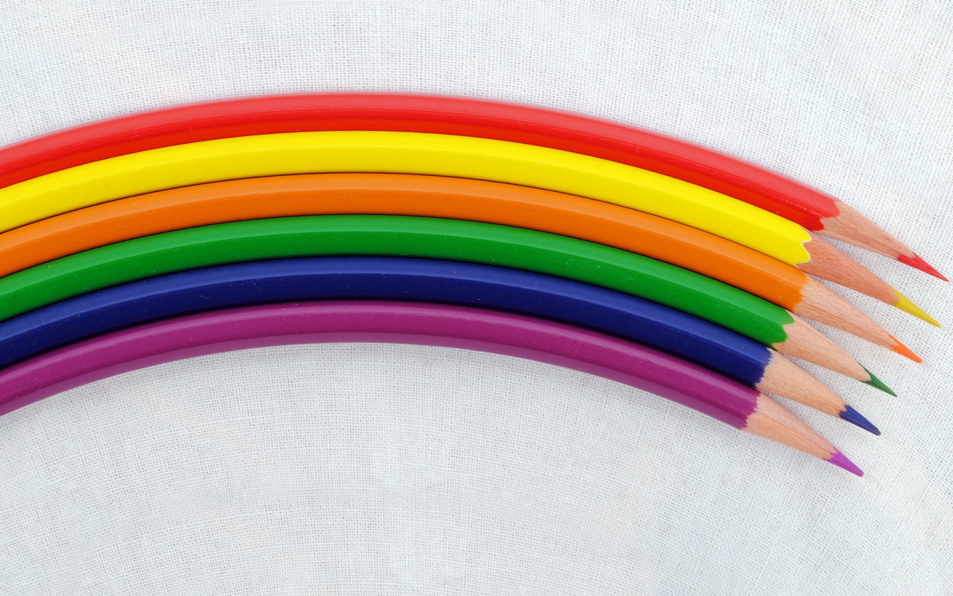 HD PC desktop wallpaper featuring a close-up photograph of seven colored pencils arranged in a rainbow arc on a light background.