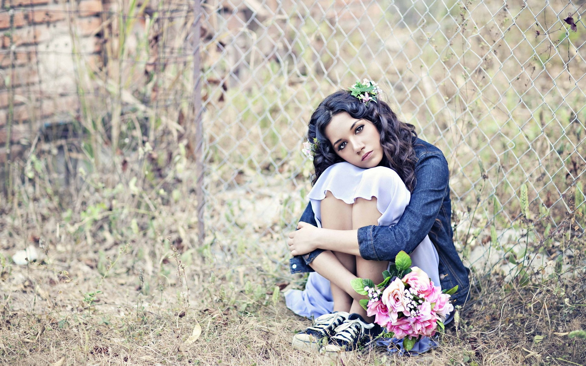 A woman sits thoughtfully among overgrown grass, holding a bouquet of pink flowers, with a serene expression, surrounded by a rustic fence. This HD desktop wallpaper captures a reflective mood.