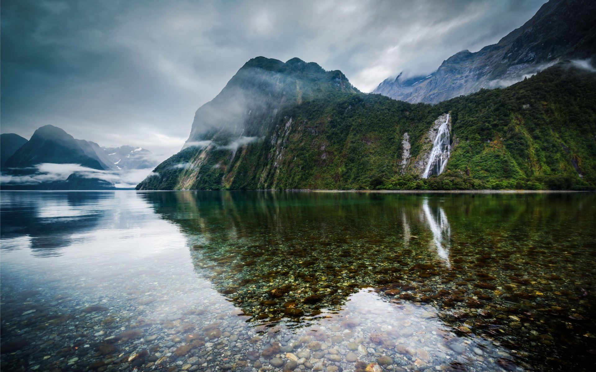 HD desktop wallpaper of a serene New Zealand mountain lake with clear stones, a cascading waterfall, and dramatic clouds reflected in the calm water.