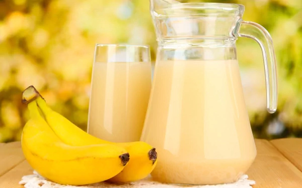 HD desktop wallpaper showing a glass and pitcher of banana smoothie with two ripe bananas on a wooden surface against a blurred yellow-green background.