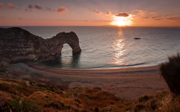 Sunset over Durdle Door limestone arch and sandy bay, cliffs and grassy foreground, sun reflecting on calm sea — 2K Quad HD PC desktop wallpaper background