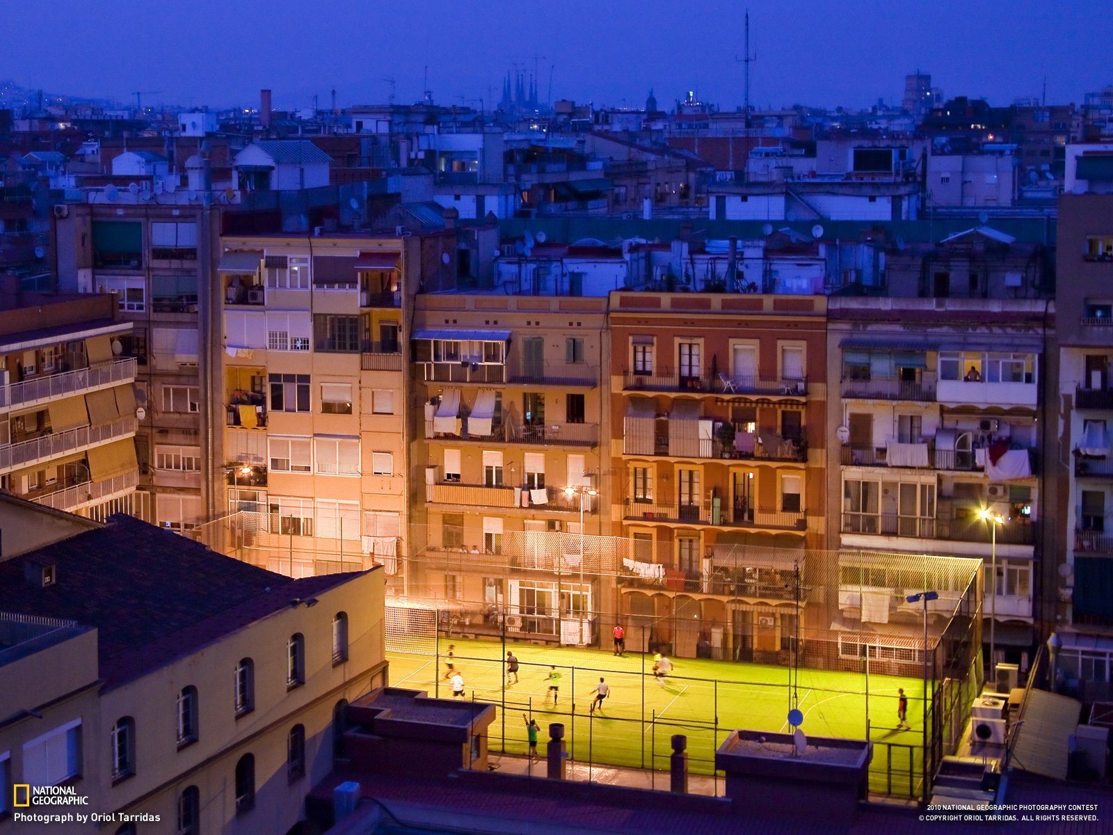 HD desktop wallpaper showing a man-made urban scene in Barcelona at dusk, featuring illuminated residential buildings and a brightly lit sports court.