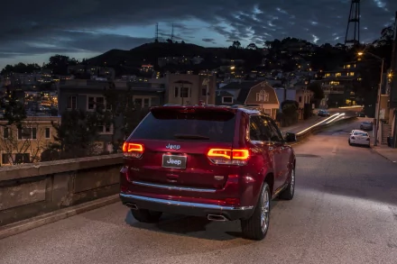 Red Jeep Grand Cherokee parked on a quiet city street at dusk, city lights and hills in the background. HD PC desktop wallpaper and background.