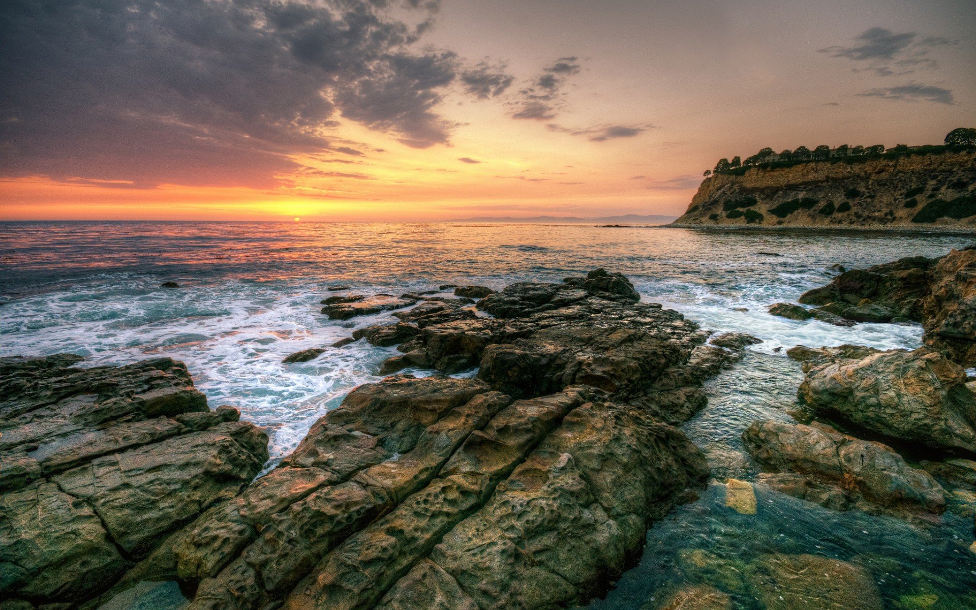 HD PC desktop wallpaper and background — nature coastline at sunset: rocky shore and tide pools, waves breaking beneath dramatic clouds and a distant cliff.