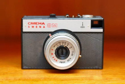 Vintage Smena 8M film camera on a warm wooden surface — 2K Quad HD PC desktop wallpaper and background; man-made camera.