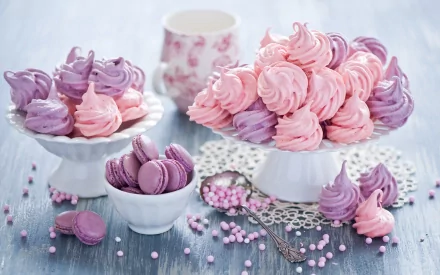 HD desktop wallpaper of colorful macarons and meringues in pastel pink and lavender shades, displayed in elegant white bowls with scattered candy beads on a light blue wooden table background.