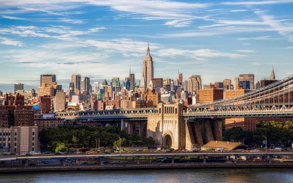 The image showcases the Manhattan Bridge with the iconic New York City skyline in the background, featuring the Empire State Building amidst a vibrant, urban landscape.