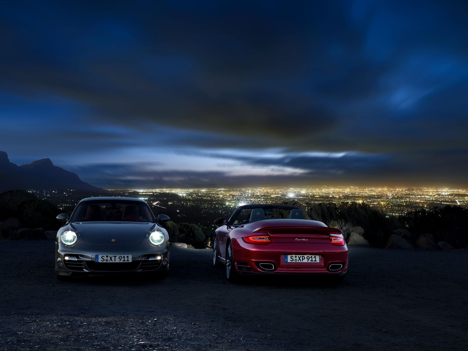HD PC desktop wallpaper: two Porsche 911s parked on a hill at night, headlights on, overlooking a glowing cityscape beneath a dramatic cloudy sky.