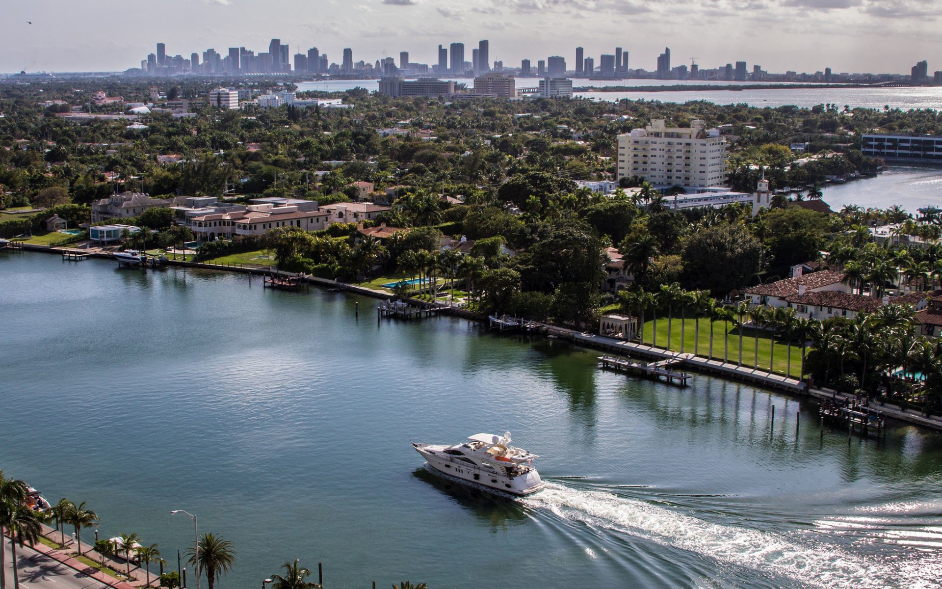 HD PC desktop wallpaper: aerial view of a Florida man-made waterfront town, a yacht cutting through a blue canal, palm-lined shores and a distant city skyline.
