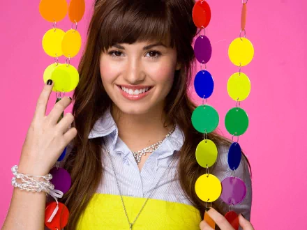 A vibrant HD desktop wallpaper featuring Demi Lovato smiling brightly with colorful hanging decorations against a pink background, capturing a lively and energetic mood.