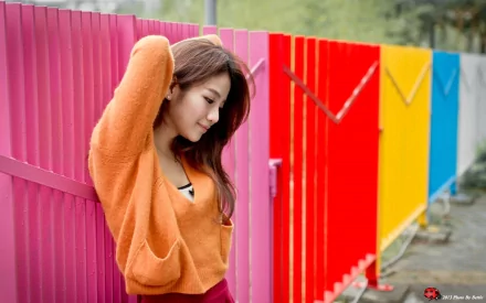 Asian woman with long hair, wearing an orange sweater, standing against a multicolored fence. The vibrant fence features panels in pink, red, yellow, and blue. HD desktop wallpaper background.