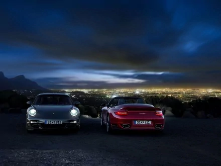 HD PC desktop wallpaper: two Porsche 911s parked on a hill at night, headlights on, overlooking a glowing cityscape beneath a dramatic cloudy sky.