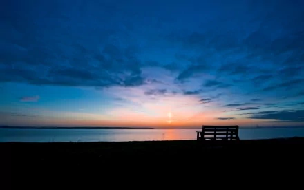 HD desktop wallpaper featuring a man-made bench silhouetted against a vibrant sunset over calm waters under a dramatic sky.