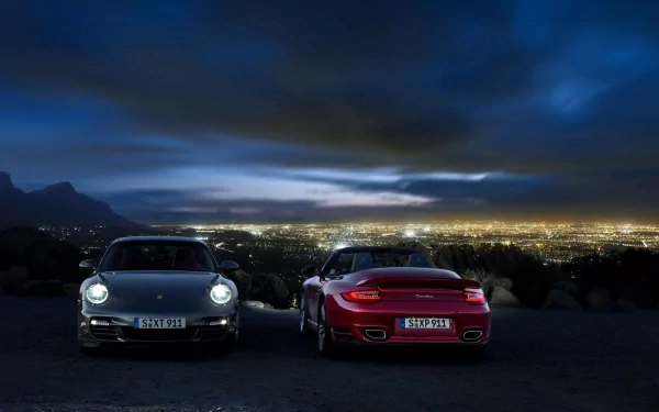 HD PC desktop wallpaper: two Porsche 911s parked on a hill at night, headlights on, overlooking a glowing cityscape beneath a dramatic cloudy sky.