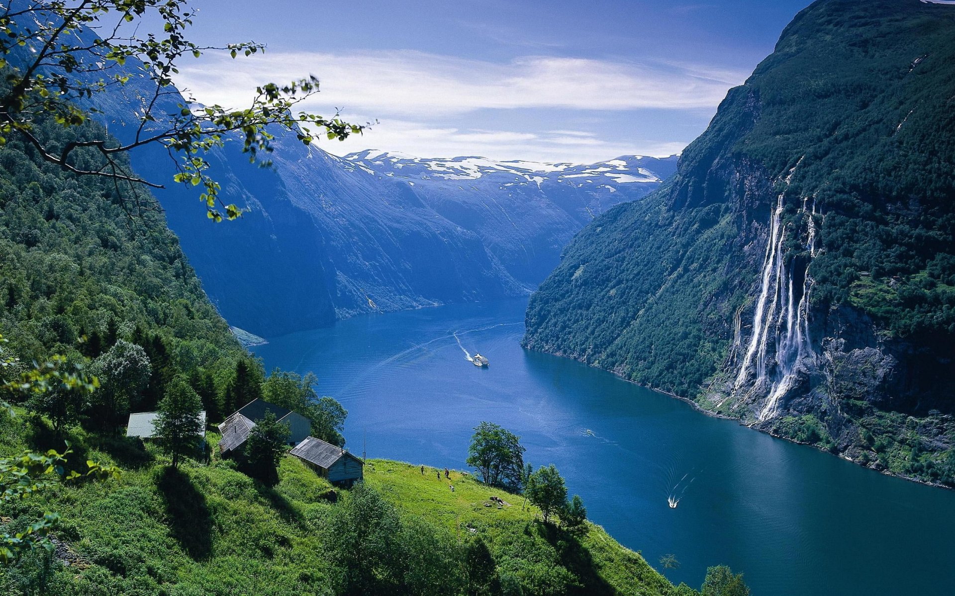 HD desktop wallpaper showing a breathtaking landscape with green mountains, a serene blue fjord, a distant waterfall, and small houses nestled on a grassy slope.
