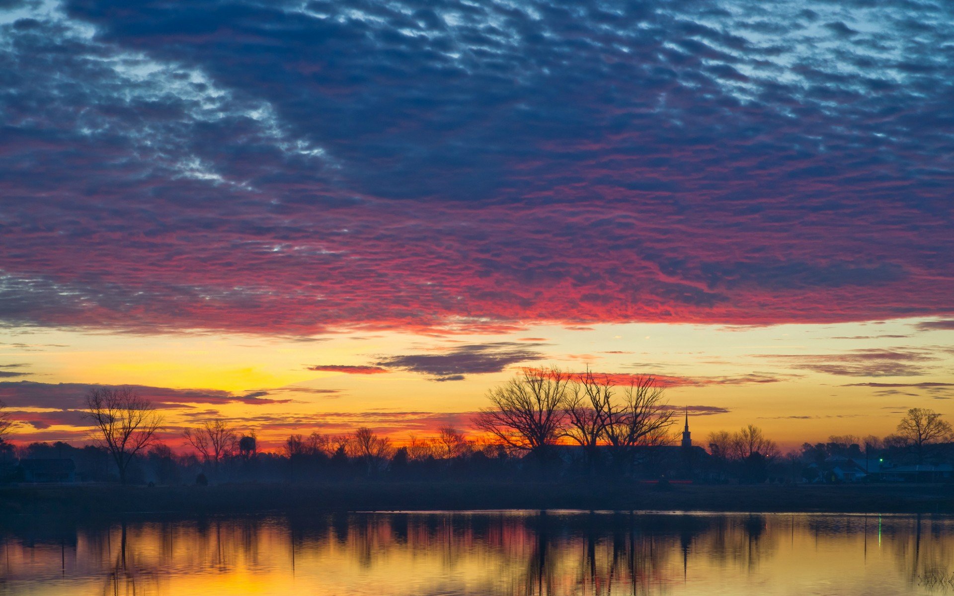 HD PC desktop wallpaper showing a vibrant sunset over a calm body of water with silhouetted trees and a textured sky blending orange, purple, and blue hues.