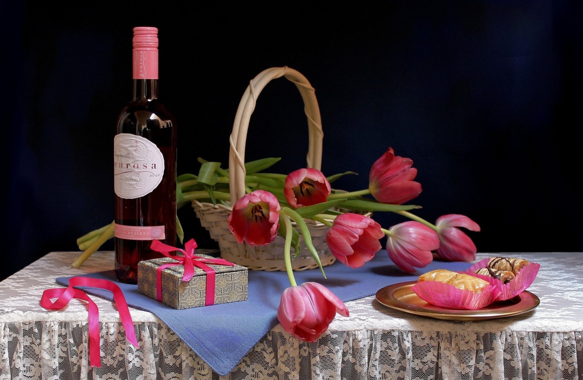 Still life featuring a bottle of wine, a basket of tulips, a gift box with a ribbon, and colorful sweets on a lace tablecloth, creating a vibrant and inviting atmosphere.