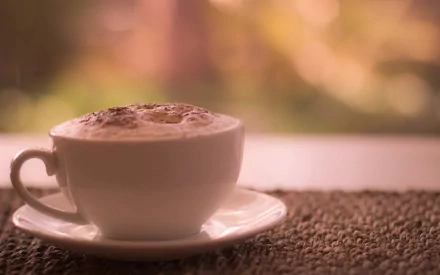 A cozy cup of coffee sits on a saucer, with a softly blurred background, creating a warm, inviting atmosphere for a desktop wallpaper.