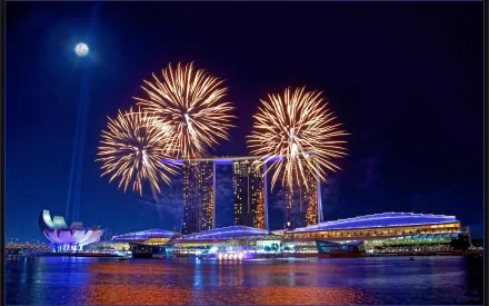 HD desktop wallpaper showcasing vibrant fireworks illuminating the night sky over a city skyline with a full moon and reflective waterfront.