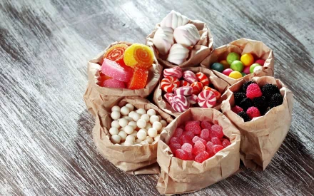 HD desktop wallpaper featuring various colorful candies in small brown paper bags arranged on a rustic wooden surface.