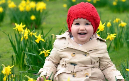 A happy child in a red knit hat and beige coat sits among blooming yellow daffodils on a green field. HD photography desktop wallpaper and background.