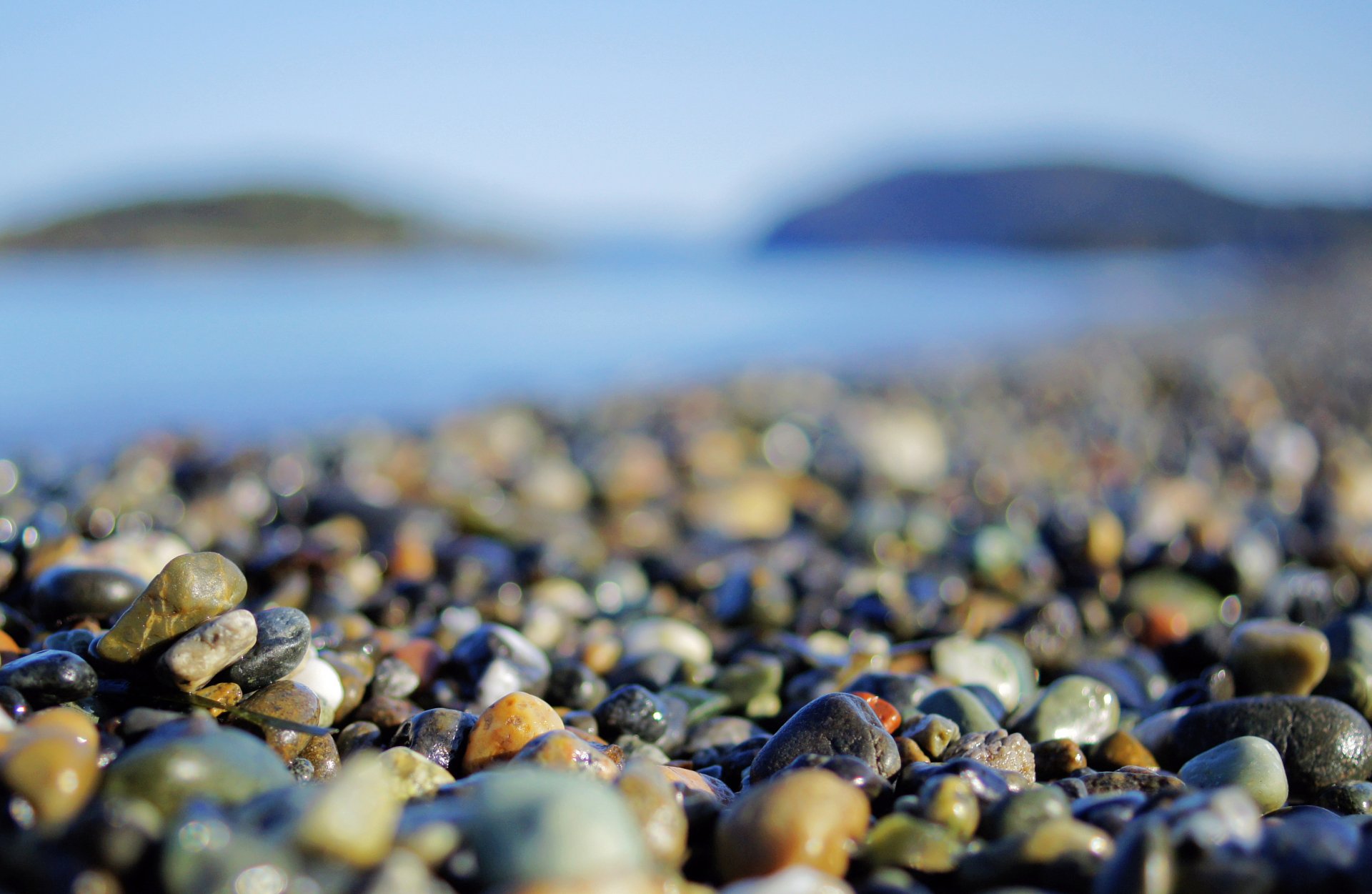 HD PC desktop wallpaper featuring a close-up of colorful stones on a beach with blurred water and distant islands in the background, highlighting nature's details.