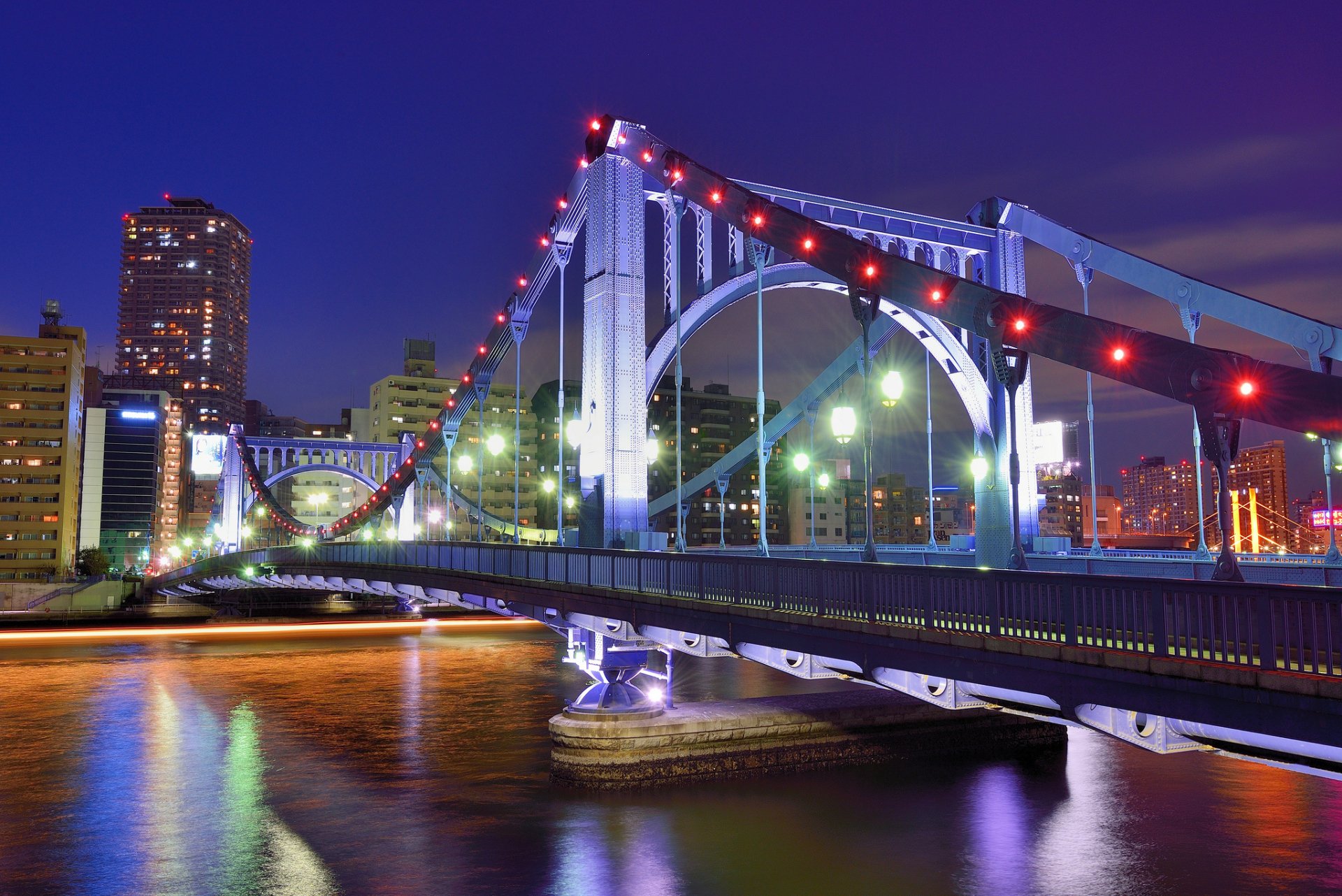 HD desktop wallpaper showing a brightly lit man-made bridge spanning a calm river against a city skyline at night.