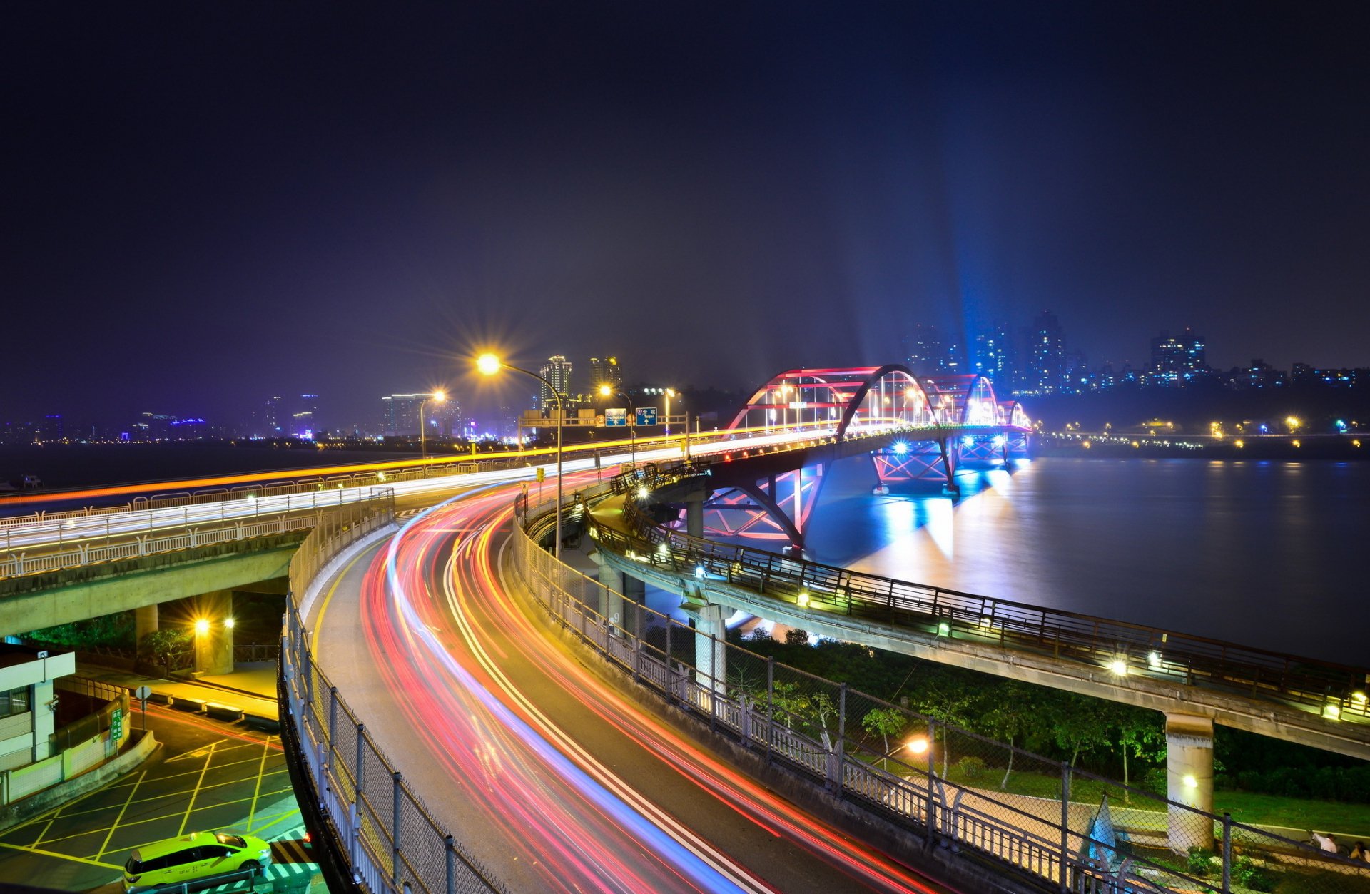 HD PC desktop wallpaper featuring a time-lapse photography of a curved bridge illuminated with colorful light trails against a nighttime urban skyline.
