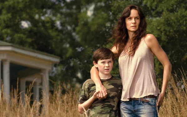 Carl Grimes, portrayed by Chandler Riggs, stands beside his mother, Lori Grimes, played by Sarah Wayne Callies, set against a serene backdrop from the TV show The Walking Dead.