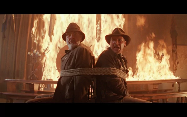 HD desktop wallpaper featuring a dramatic scene from the movie Indiana Jones and the Last Crusade with two characters tied back-to-back amid flames.
