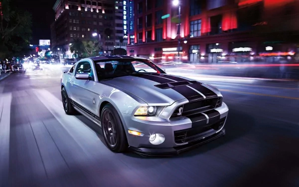 HD desktop wallpaper of a Ford Mustang speeding through a brightly lit city street at night, with vibrant lights and buildings in the background.