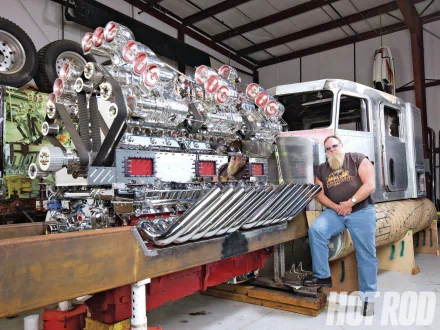 HD PC desktop wallpaper: Peterbilt vehicle in a workshop — gleaming chrome engine pipes and a bearded man seated on the bumper.