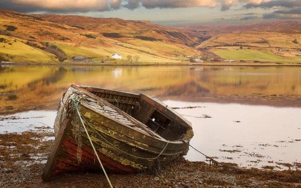 HD desktop wallpaper showing an old wooden boat resting on a calm shore with rolling hills and cloudy sky in the background.