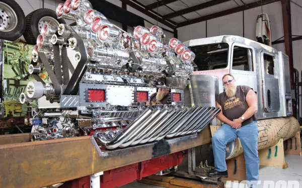 HD PC desktop wallpaper: Peterbilt vehicle in a workshop — gleaming chrome engine pipes and a bearded man seated on the bumper.