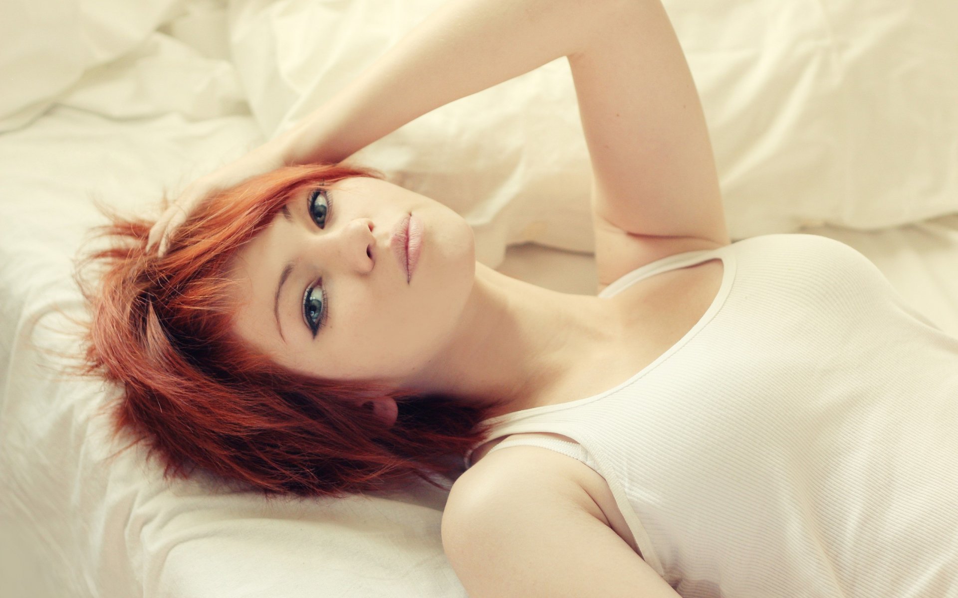 HD desktop wallpaper featuring a woman model lying on a bed, gazing upwards. She has short red hair and is wearing a white tank top, set against a soft white background.