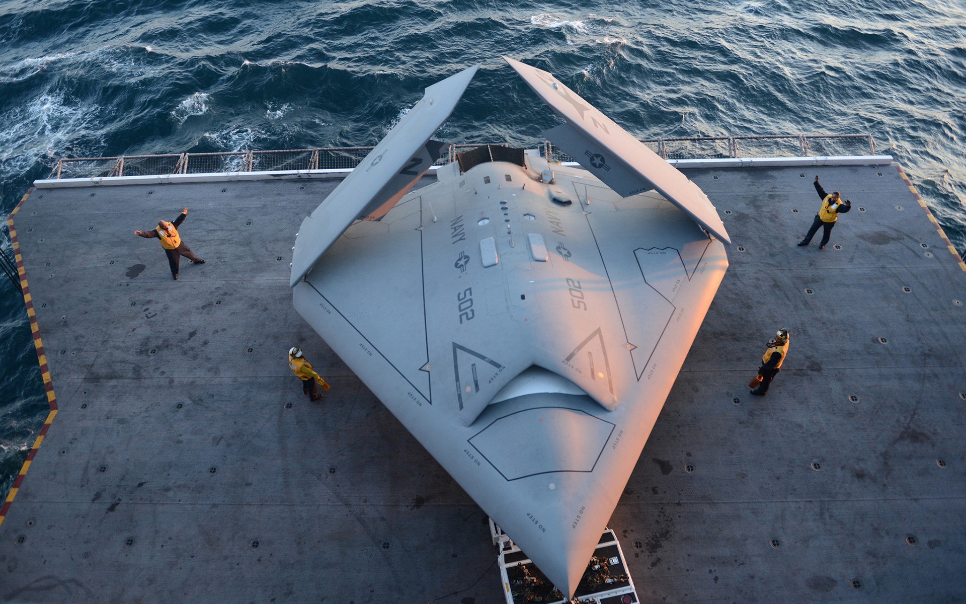 HD wallpaper of a UAV on a ship deck with crew members, set against an ocean backdrop at sunset.