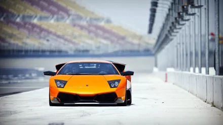 A vibrant orange Lamborghini Murcielago parked on a racetrack, showcasing its sleek design and powerful stance, set against a backdrop of empty spectator stands.