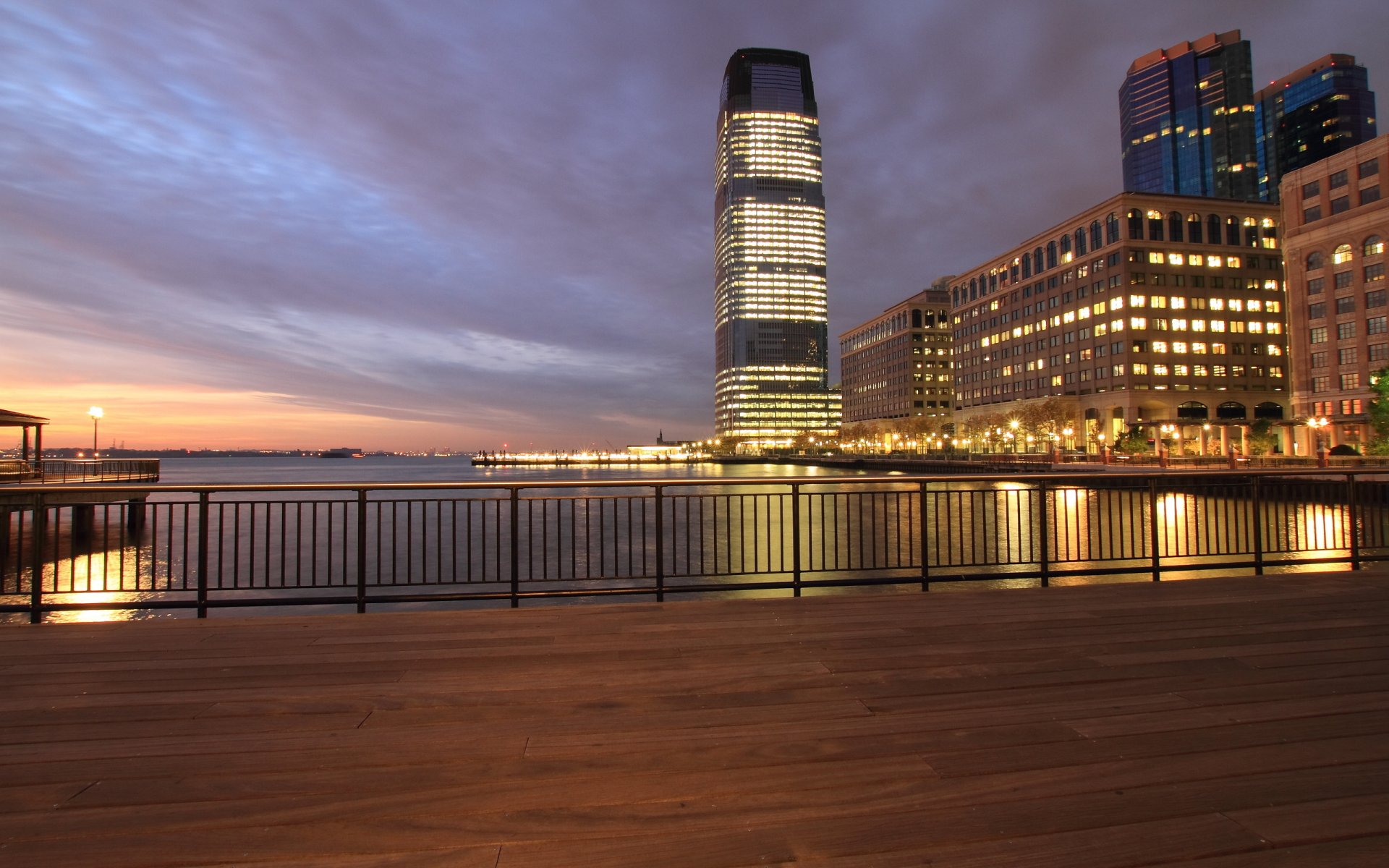A stunning HD wallpaper showcasing a waterfront view in Jersey City at dusk, featuring a modern building illuminated against a cloudy sky and reflections in the water.