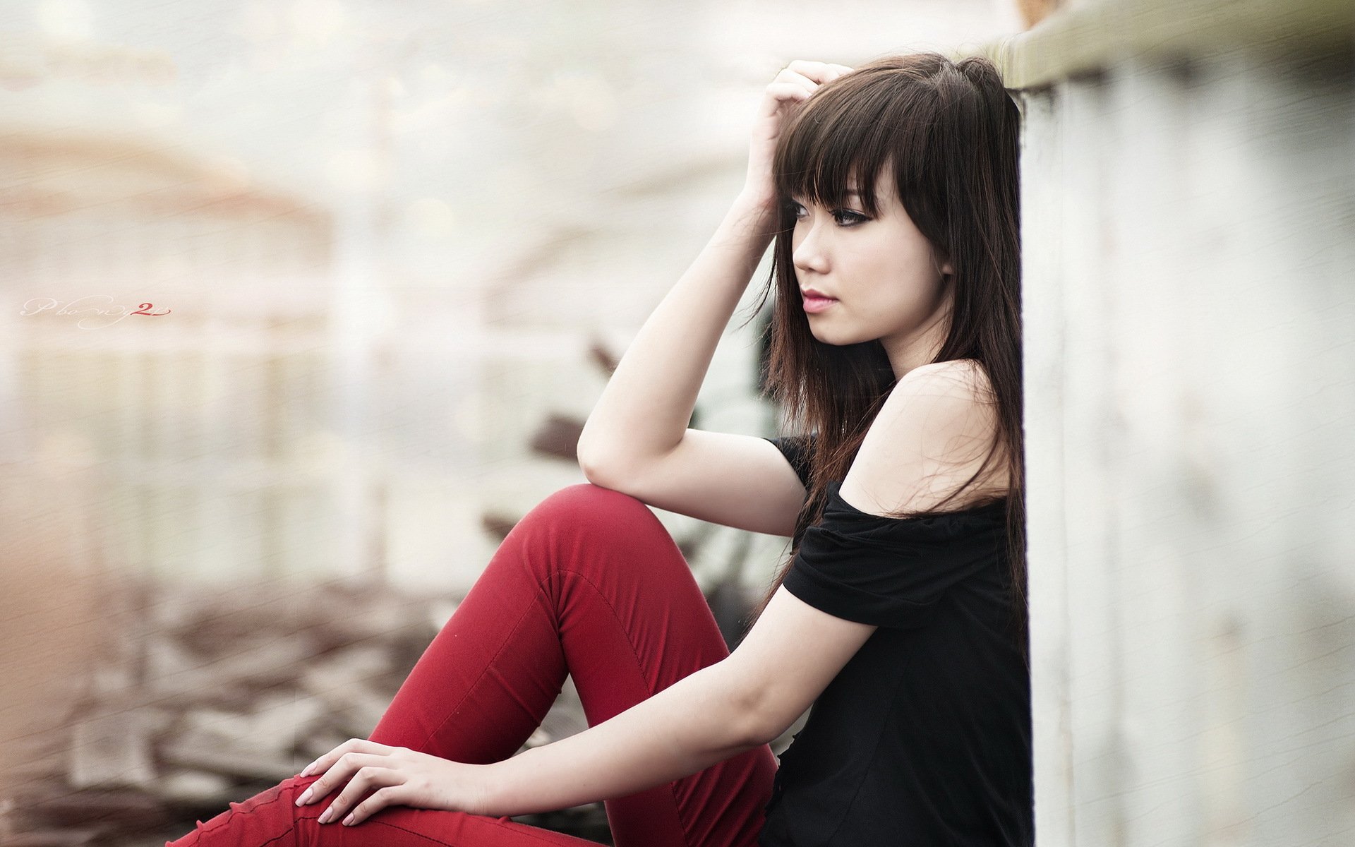 A serene portrait of an Asian woman sitting against a wall, wearing a black off-shoulder top and red pants, surrounded by soft lighting, making it a striking HD desktop wallpaper.